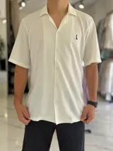 Camisa de manga corta color blanco marfil, confeccionada en tejido texturizado con cuello camisero, cierre frontal de botones y un pequeño bordado oscuro en el pecho.