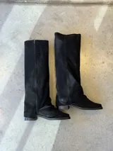 Bota de caña alta confeccionada en gamuza negra, con diseño de silueta holgada y cierre lateral completo. Presenta punta cuadrada y taco bajo cuadrado.