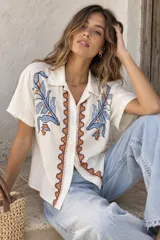 Camisa de manga corta color beige, con cuello camisero, cierre frontal con botones y bordados decorativos en azul y naranja en la parte delantera.