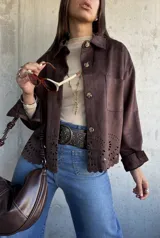 Chaqueta de gamuza color chocolate, con corte recto, cuello clásico y cierre frontal con botones. Presenta un diseño calado decorativo en el ruedo y bolsillos frontales tipo parche.