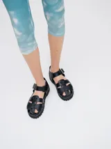 Sandalias de cuero marrón con tiras entrecruzadas, suela de goma negra con pespuntes blancos y hebilla lateral.