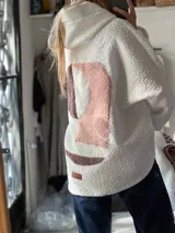 Campera con capucha confeccionada en tejido tipo corderito blanco, con diseño de collage abstracto en la espalda en tonos rosado y marrón. Presenta un corte holgado y una etiqueta de cuero sintético en la parte inferior.