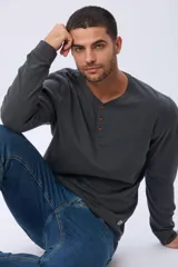 Remera de algodón texturada color gris oscuro, de manga larga, con cuello redondo y botones en la parte frontal. Presenta un pequeño logo bordado en el pecho y un parche decorativo en la manga izquierda.