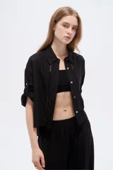Camisa negra de viscosa, holgada, con mangas cortas abotonadas, cuello camisero, detalles de cintas decorativas verticales y cierre frontal con botones.