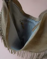 Bolso tipo hobo de gran tamaño en color marrón, confeccionado en gamuza. Presenta un diseño con flecos largos en la parte inferior, cierre superior y una correa trenzada con detalles artesanales.