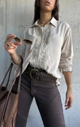 Camisa de color beige con diseño romántico, presenta una pechera central con encaje floral y botones frontales. Posee mangas largas con puños decorados con volados y encaje a juego.