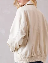 Campera bomber de eco cuero color beige, con cuello camisero, cierre frontal metálico y bolsillos delanteros con solapa y botón a presión. Presenta mangas largas con puños abotonados y ruedo trasero elastizado.