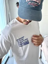 Remera blanca de manga corta con estampado gráfico en azul en la espalda, que incluye el texto "BROKERS GRAPHIC RESEARCHING" y varios logos de la marca.