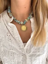 Collar tipo gargantilla compuesto por piedras naturales en tonos celestes y verdes con separadores dorados, presenta un dije central grande en forma de sol de metal dorado.