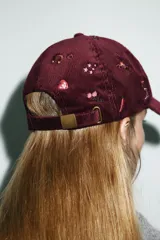 Gorra tipo baseball confeccionada en pana color bordó, con múltiples bordados decorativos de motivos variados como cactus, caballos, corazones y estrellas. Presenta ajuste posterior y visera curva.