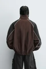 Cazadora de ajuste relajado, color marrón oscuro con bloques de color negro en las mangas. Presenta ribetes blancos que delinean los paneles y cuello alto con cierre frontal de cremallera. Los puños y el bajo son elásticos.