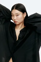 Blusa negra oversized en tejido liviano, con cuello clásico, tapeta con botones ocultos, canesú doble con frunces en el delantero y la espalda, y mangas globo largas con puños abotonados.