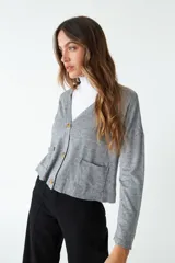 Cardigan de tejido de punto color gris melange, con escote en V, cierre frontal con botones y bolsillos tipo cartera. Presenta un diseño de corte recto y mangas largas.