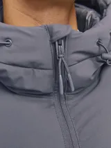 Campera puffer con capucha ajustable, cierre frontal con cremallera y bolsillos laterales. Presenta un diseño acolchado en color gris oscuro con logo sutil bordado en el pecho.