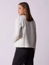 Chaqueta de tejido tweed color blanco con destellos brillantes, de corte recto y estructurado. Presenta cuello a la base, abertura frontal y puños decorados con botones en contraste. Los bordes del cuello, la cartera frontal y el ruedo cuentan con una terminación deshilachada.