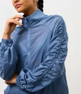 Campera deportiva tipo parka de microfibra en color azul, con cuello alto, cierre frontal por cremallera y mangas largas con puños elásticos. Presenta un diseño con fruncido lateral ajustable y corte amplio.
