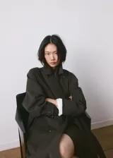 Chaqueta estilo gabardina larga en color gris oscuro, con doble botonadura frontal, solapas clásicas, bolsillos y trabillas ajustables en los puños.