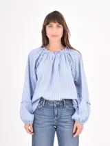 Blusa de algodón a rayas verticales en tonos azul claro y blanco, con cuello redondo fruncido y lazo de ajuste. Presenta mangas largas abullonadas con detalle de volados en los hombros y puños elásticos.