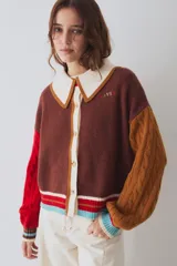 Cardigan de punto multicolor con diseño colorblock, cuello polo rosado con borde rojo, cierre frontal de botones, mangas largas abuchonadas (una roja y otra verde militar con tejido de ochos) y detalles de rayas en los puños y el ruedo.