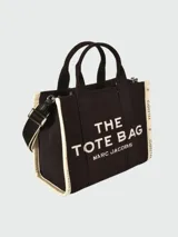 Bolso tote Marc Jacobs modelo The Jacquard, confeccionado en tejido de lona negro con detalles en blanco. Presenta el logo de la marca y la inscripción 'THE TOTE BAG' en el frente, con doble asa superior y correa ajustable extraíble.