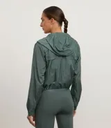 Campera cortavientos deportiva de corte cropped, color verde oscuro. Presenta capucha, cierre frontal con cremallera, mangas largas con puños elásticos y cordón ajustable en el ruedo.
