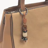 Cartera tipo tote de color khaki y marrón, con diseño combinado de textura lisa y tejido tipo sarga. Incluye doble asa de mano, correa larga extraíble y un dije decorativo con cuentas de madera y piedra colgando de una de las asas.