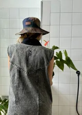 Vestido corto de jean color gris oscuro con efecto acid wash, sin mangas, con cuello camisero y cierre frontal de botones. Incluye dos bolsillos en el pecho con solapa y pespuntes decorativos.