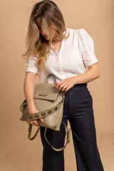 Mochila tipo cartera con tapa, confeccionada en símil cuero color suela. Cuenta con doble compartimento, división interna, cierre de seguridad y bolsillo trasero con cierre. Incluye una correa frontal decorativa con tachas metálicas.