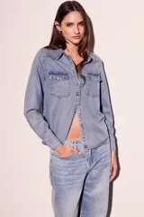 Camisa de denim celeste con cuello clásico, cierre frontal con botones a presión, dos bolsillos con solapa abotonada en el pecho y mangas largas con puños abotonados.