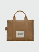 Bolso tipo tote de lona color negro con letras blancas bordadas en el frente que dicen 'THE TOTE BAG MARC JACOBS'. Cuenta con asas superiores cortas y una correa de hombro ajustable y extraíble.