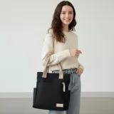 Tote bag de lona color negro con asas y detalles en color beige. Presenta un compartimento principal, un bolsillo frontal con cierre de cremallera, bolsillos laterales abiertos y un parche decorativo en la parte inferior.