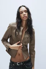 Blazer corto de estilo militar color tostado, con cuello redondo, manga larga y cierre frontal con ganchos metálicos. Presenta detalle de botones decorativos dorados en el frente y en los hombros, con costuras marcadas y forro interior a contraste.