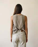 Blusa sin mangas de diseño fluido, confeccionada en tejido gris con rayas verticales finas. Presenta cuello redondo, cierre frontal con botones y detalles de volados escalonados en los laterales con terminaciones en puntilla blanca.