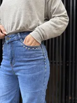 Pantalón de jean azul de corte recto y tiro alto, con detalle de tachas metálicas en los bolsillos delanteros y cierre frontal con botón.