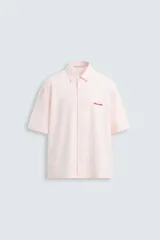 Camisa de algodón color rosa pálido, corte boxy fit, con cuello solapa, manga corta y abotonadura frontal. Presenta un texto bordado en contraste de color rojo en el delantero.