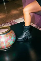 Bota de caña media confeccionada en cuero negro, con suela gruesa track antideslizante. Presenta un diseño estilo biker con estribo integrado, hebillas metálicas decorativas y una correa ajustable en la parte superior de la caña. Incluye cierre lateral para facilitar el calce.