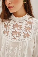 Camisa blanca de estilo romántico con cuello alto, confeccionada en algodón con detalles de encaje y bordados en el frente. Presenta mangas largas abullonadas con puños elásticos y terminaciones de encaje.