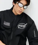 Campera bomber negra de microfibra, con cierre frontal por cremallera, puños y ruedo elásticos, y bolsillos con cierre. Cuenta con múltiples parches y estampas bordadas alusivas a la NASA, incluyendo el logo principal en el pecho, insignias en la manga derecha y una bandera de Estados Unidos.