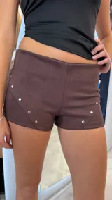 Short de gamuza color marrón chocolate, con diseño de tiro bajo y detalle de costuras decorativas con tachas metálicas plateadas en los laterales.