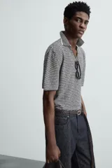 Polo de punto de manga corta con cuello solapa y abertura frontal en forma de V. Presenta un patrón de rayas horizontales finas en blanco y negro, con un acabado texturizado y un calce relajado.