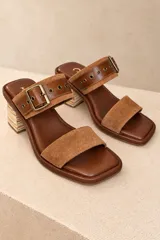 Sandalias de cuero marrón, con taco cuadrado de madera, punta cuadrada y dos tiras superiores. La tira superior cuenta con una hebilla metálica grande y ojalillos decorativos.