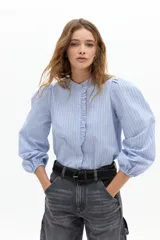 Camisa de algodón a rayas verticales celestes y blancas, con cuello mao, mangas largas abullonadas con puños elásticos y detalle de volado en la cartera de botones frontal.