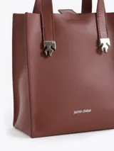 Cartera tipo tote pequeña de cuero color tostado, con doble asa superior y herrajes metálicos plateados en forma de llama en la base de las manijas. Presenta el logo de la marca estampado en la parte inferior frontal.