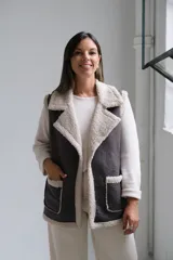 Chaleco de gamuza sintética en color gris oscuro con interior y bordes de corderito beige. Presenta solapas amplias, dos bolsillos frontales con ribetes de corderito y diseño sin cierre.