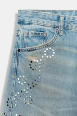 Jeans de tiro bajo y corte wide leg, confeccionados en denim de color celeste claro con lavado desgastado. Presentan detalles de roturas y aplicación de tachas metálicas en los laterales y bolsillos delanteros. Cierre frontal con cremallera y botón.
