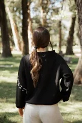Buzo de punto color negro con diseño de bordado en hilo crudo que forma líneas curvas decorativas. Presenta cuello redondo, mangas largas con caída relajada y corte holgado.