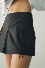 Falda pantalón negra de tiro medio, confeccionada en algodón, con pliegue frontal y maxi bolsillos cargo con solapa y cremallera en ambos lados.