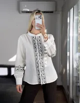 Camisa de manga larga color blanco con bordados geométricos en negro a lo largo de la botonadura frontal y en los puños. Presenta cuello mao y cierre frontal con botones.