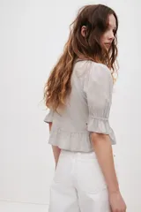 Blusa color gris claro en tejido de mezcla de viscosa y algodón, con escote en V pronunciado, volados frontales, alforzas, inserciones de encaje y detalles bordados. Cuenta con mangas abullonadas al codo con puños elásticos de volado y volado ancho en el bajo.