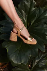 Sandalias de taco cuadrado medio, con base y talón en gamuza color tostado. Presenta una tira frontal de múltiples bandas doradas y una pulsera al tobillo con hebilla metálica.